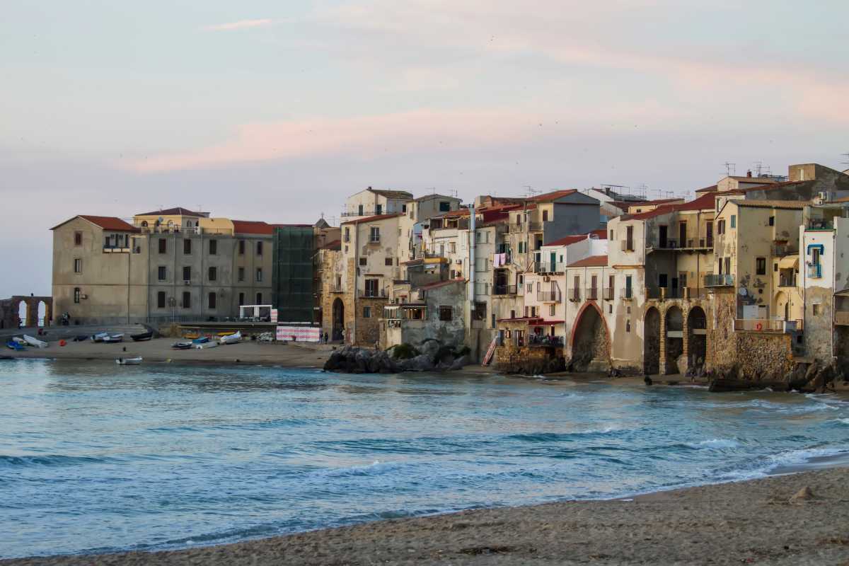 Una famiglia con bambini piccoli che gioca sulla spiaggia dorata di Cefalù, con la Rocca e il borgo sullo sfondo, atmosfera gioiosa e rilassata