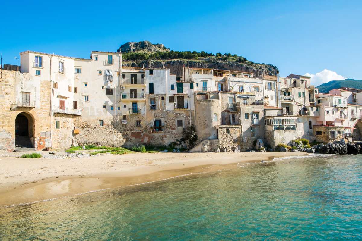 Panorama della spiaggia di Cefalù all’alba con la Rocca sullo sfondo, barche di pescatori sulla riva e le prime luci dorate che illuminano il borgo antico